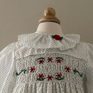 Op de afbeelding: Witte smockjurk met zwarte stippen en rode geborduurde bloemen. De jurk heeft een witte geruite kraag met een rode bloemenaccent.