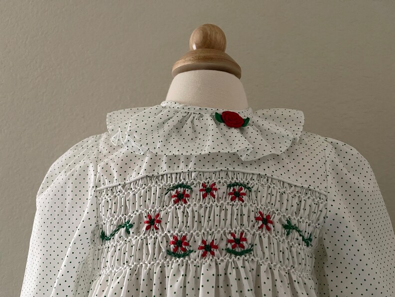 Op de afbeelding: Witte smockjurk met zwarte stippen en rode geborduurde bloemen. De jurk heeft een witte geruite kraag met een rode bloemenaccent.