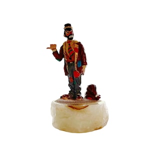 May include: A hand-painted figurine of a clown with a red nose, black hair, and a brown hat. The clown is wearing a red, brown, and blue patched coat and is holding a bag. The figurine is standing on a white marble base.