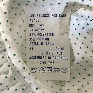 Op de afbeelding: Wit shirt met groene stippen en een wit label met wasinstructies. Het label zegt "See reverse for care", "78435", "BNS 5/45", "RN 40672", "65% Polyester", "35% Katoen", "Style H 481 S", "29", "AH 91", "U.S. Materials", "Assembled in Barbados", "Size T-2".