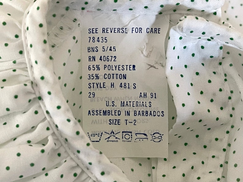 Op de afbeelding: Wit shirt met groene stippen en een wit label met wasinstructies. Het label zegt "See reverse for care", "78435", "BNS 5/45", "RN 40672", "65% Polyester", "35% Katoen", "Style H 481 S", "29", "AH 91", "U.S. Materials", "Assembled in Barbados", "Size T-2".