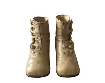 May include: A pair of vintage beige leather ankle boots. Each boot features a row of round buttons on the side and decorative perforations near the toe. The boots have a rounded toe and a short heel, showcasing a classic, timeless design.
