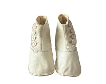 May include: A pair of off-white baby boots. Each boot features a rounded toe, a button closure on the side, and a high-top design. The boots are made of a soft, textured material and appear to be vintage or antique.