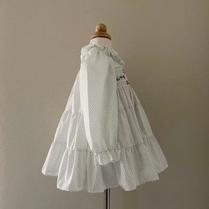 Op de afbeelding: Witte jurk met zwarte stippen en een gerimpelde halslijn. De jurk heeft een smok-lijfje en een gelaagde rok.