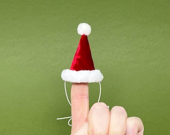 May include: A miniature Santa hat, red with white trim and a white pom-pom, is displayed. The hat is small enough to fit on a finger, suggesting a novelty or holiday-themed item. The background is a solid green.