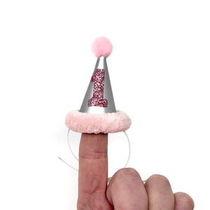 May include: A silver party hat with a pink pom-pom on top and a pink glitter number "1" on the front. The hat is worn on a finger.