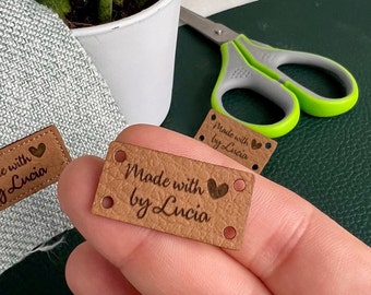 Op de afbeelding: Twee bruine leren labels met de tekst "Made with ♥ by Lucia" in een bruin lettertype. De labels hebben elk vier gaten.