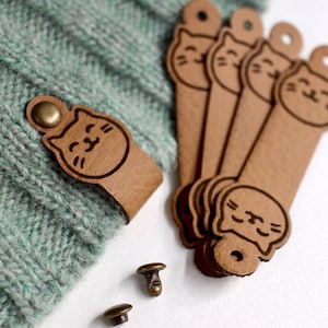 May include: Six brown leather cat-shaped toggles with a smiling face design. The toggles are attached to a green knitted fabric. Two small metal snaps are also included.