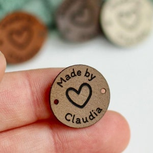 May include: A small, round, wooden tag with the text "Made by Claudia" and a heart symbol. The tag is brown with a dark brown heart and text. Other similar tags are visible in the background.