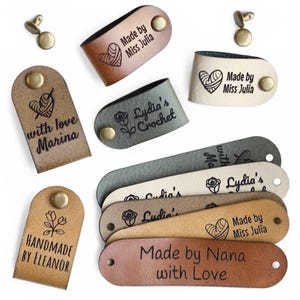 Op de afbeelding: Een verzameling leren labels in verschillende kleuren, waaronder bruin, beige en grijs. Elk label heeft tekst en een kleine afbeelding, zoals een hart of bloem, en is bevestigd met een messing klinknagel. Ontworpen voor handgemaakte ambachten.