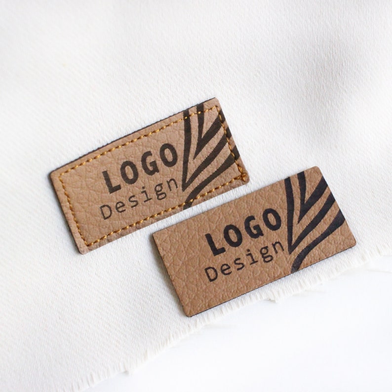 Op de afbeelding: Twee bruine lederen labels met gouden stiksels. De labels hebben een zwart-wit grafisch ontwerp en de tekst "Logo Design" erop gedrukt.