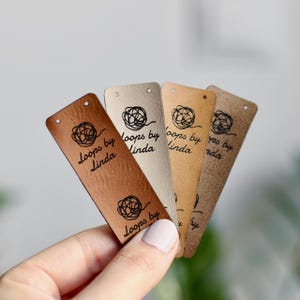 Op de afbeelding: Een verzameling leren labels in verschillende tinten bruin, beige en tan. Elk label heeft de tekst "Loops by Linda" en een gestileerd cirkelvormig ontwerp. De labels hebben een klein gaatje aan de bovenkant voor bevestiging.