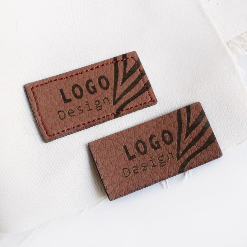 Op de afbeelding: Twee bruine leren labels met de tekst "LOGO Design" in zwarte letters. De labels hebben een gestructureerd oppervlak en een decoratief patroon.