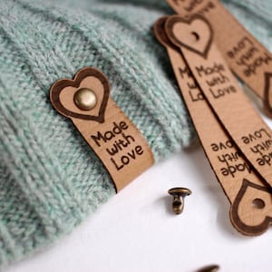 May include: Close-up of a light green knitted fabric with a brown leather tag attached. The tag has a heart shape with the text "Made with Love" engraved on it. A small metal rivet is visible on the tag.