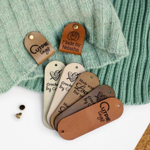 Könnte beinhalten: Sechs braune Leder-Etiketten mit verschiedenen Texten und Designs. Die Etiketten sind oval geformt und haben oben ein Loch zum Anbringen. Der Text auf den Etiketten lautet "Cutpie Tags", "Made by Natasha", "Made with Love", "Crocheted by C" und "Crocheted for you by C".