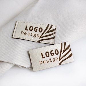 Op de afbeelding: Twee witte leren labels met bruine stiksels en een bruin grafisch ontwerp. De tekst op de labels luidt "LOGO Design".
