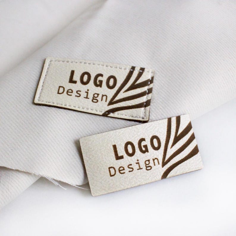 Op de afbeelding: Twee witte leren labels met bruine stiksels en een bruin grafisch ontwerp. De tekst op de labels luidt "LOGO Design".