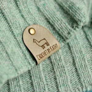 May include: A light green knitted fabric with a brown leather tag attached. The tag has a small illustration of a llama and the text "CROCHET BY KATHY".
