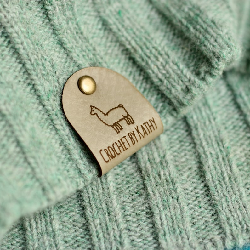 May include: A light green knitted fabric with a brown leather tag attached. The tag has a small illustration of a llama and the text "CROCHET BY KATHY".