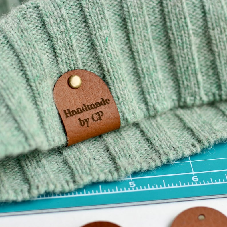 May include: A close-up of a brown leather tag with the text "Handmade by CP" on a light green knitted fabric.