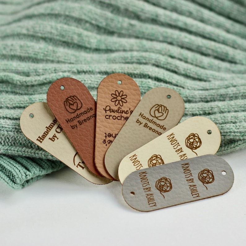 May include: Six leather tags with different designs and text. The tags are oval shaped and have a hole punched at the top. The text on the tags includes "Handmade by Breana", "Pauline's Crochet", "Knots by Ashley", and a heart with a hand on either side.