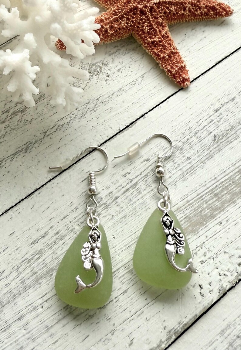 May include: A pair of earrings with green sea glass teardrop dangles and silver mermaid charms.