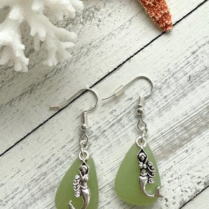 May include: A pair of green sea glass earrings with silver mermaid charms hanging from silver hooks.