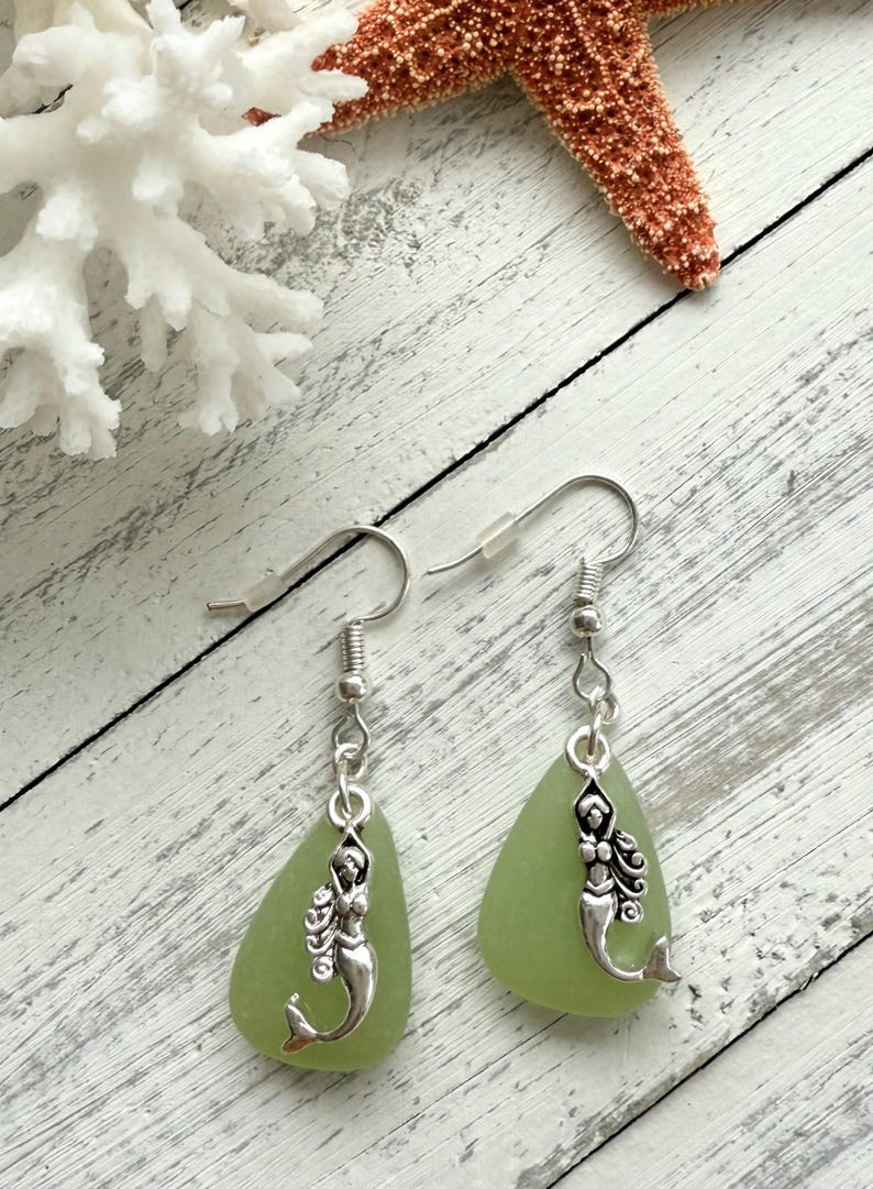 May include: A pair of green sea glass earrings with silver mermaid charms hanging from silver hooks.