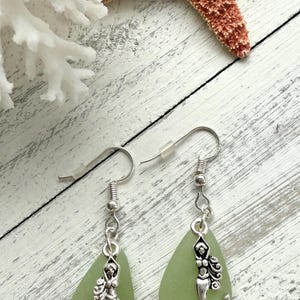May include: A pair of green sea glass earrings with silver mermaid charms. The earrings are teardrop shaped and dangle from silver hooks.
