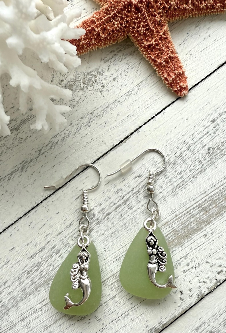 May include: A pair of green sea glass earrings with silver mermaid charms. The earrings are teardrop shaped and dangle from silver hooks.