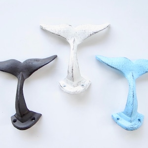May include: Three whale tail hooks in different colors: black, white, and light blue. The hooks are made of cast iron and have a rustic finish.