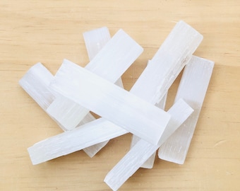 May include: A group of white selenite sticks, also known as selenite wands, on a light brown wooden surface. The sticks are smooth and have a natural, crystalline texture.