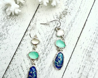 May include: Dangle earrings with a silver finish, featuring three colorful stones: a white oval, a light green teardrop, and a dark blue, glittery oval. The earrings are displayed on a white wooden surface.