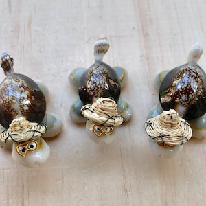 May include: Three small turtle figurines made from seashells. Each turtle is wearing a straw hat and has a pair of gold-rimmed glasses.