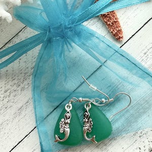 May include: A pair of green sea glass earrings with silver mermaid charms. The earrings are packaged in a light blue organza bag.