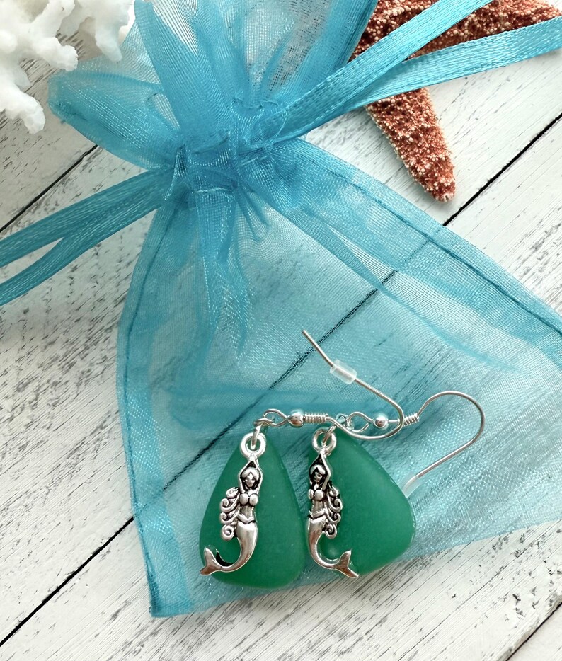 May include: A pair of green sea glass earrings with silver mermaid charms. The earrings are packaged in a light blue organza bag.