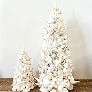 May include: Two white seashell Christmas trees, one large and one small, on a wooden surface. The trees are made entirely of white seashells of various sizes and shapes.