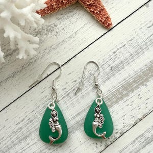 May include: A pair of dangle earrings featuring a mermaid charm on a teardrop-shaped piece of sea glass. The sea glass is a vibrant green color.