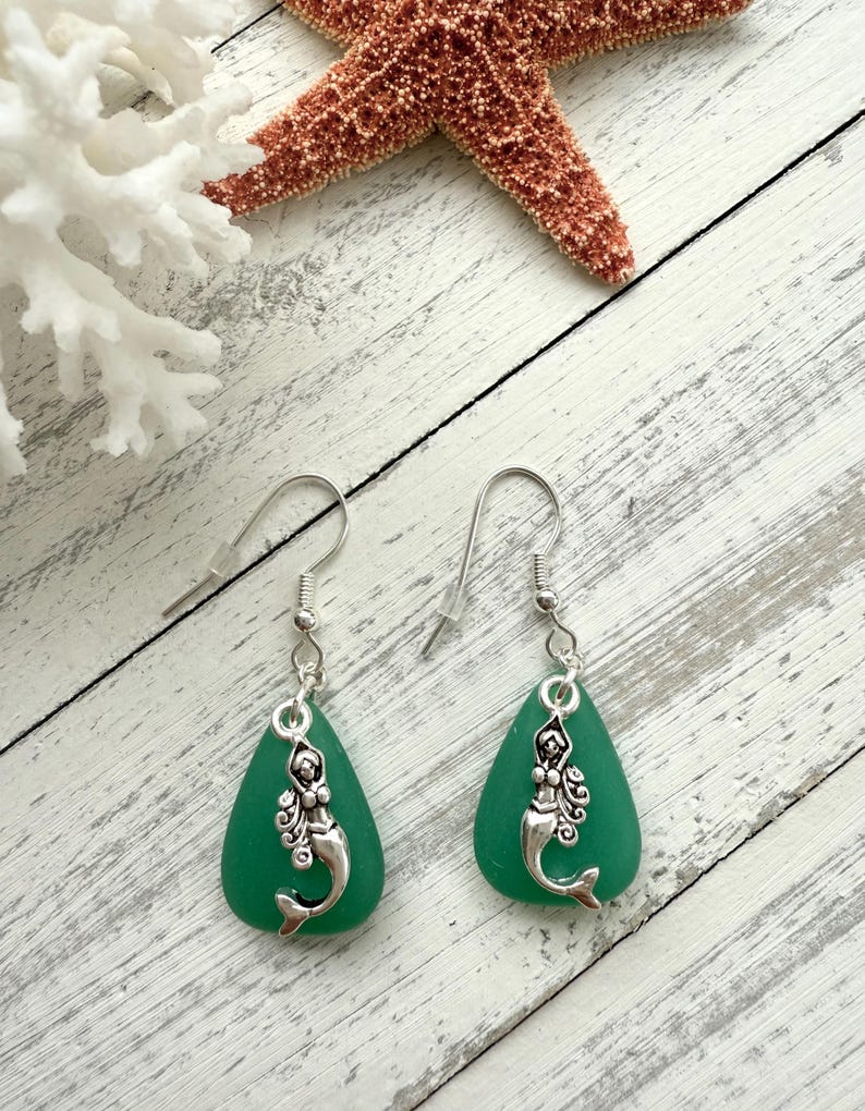 May include: A pair of dangle earrings featuring a mermaid charm on a teardrop-shaped piece of sea glass. The sea glass is a vibrant green color.