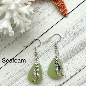 May include: Seafoam green glass earrings with silver mermaid charms. The earrings are on a white wooden surface with a white coral and a starfish.