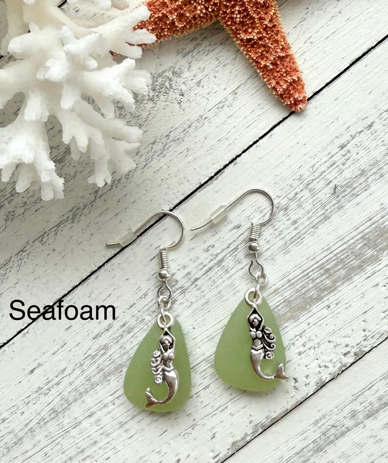 May include: Seafoam green glass earrings with silver mermaid charms. The earrings are on a white wooden surface with a white coral and a starfish.