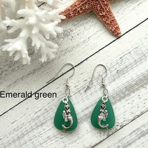 May include: A pair of emerald green sea glass earrings with silver mermaid charms hanging from silver hooks.