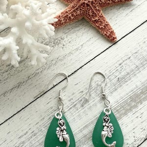 May include: A pair of green sea glass earrings with silver mermaid charms. The earrings are teardrop shaped and have silver hooks.