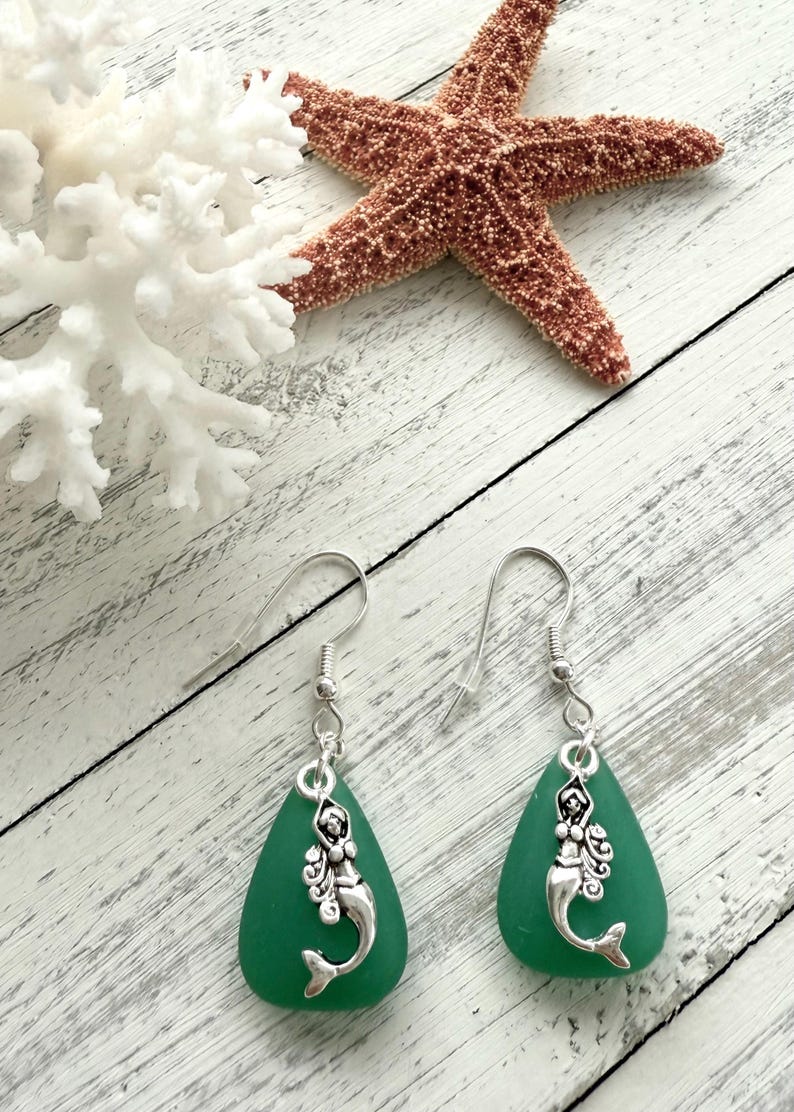 May include: A pair of green sea glass earrings with silver mermaid charms. The earrings are teardrop shaped and have silver hooks.