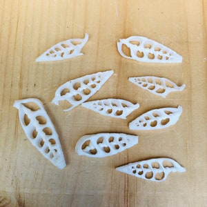 May include: Eight white, decorative, laser-cut, leaf-shaped pieces with intricate designs. The pieces are arranged on a light brown wooden surface.