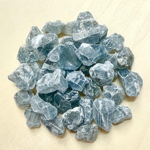 May include: A pile of rough, blue-gray crystals with irregular shapes. The crystals have a translucent quality, and the light reflects off their textured surfaces. The crystals are arranged in a circular pattern against a light-colored background.