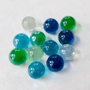 May include: A collection of glass marbles in various shades of blue, green, and clear. The marbles are round and appear to be made of transparent glass, with some reflecting light. The marbles are scattered on a white surface.