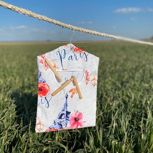 May include: A white fabric garment bag with a Paris theme, featuring the Eiffel Tower, pink flowers, and the word "Paris" in blue script. Two wooden clothespins are attached. The bag hangs from a metal hanger.