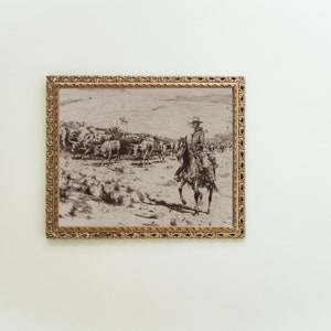 May include: A black and white framed print depicting a cowboy riding a horse through a herd of cattle.