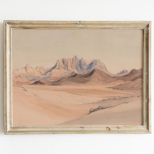 May include: A framed watercolor painting depicting a desert landscape with a mountain range in the background. The mountains are a light blue color, and the desert is a light brown color.